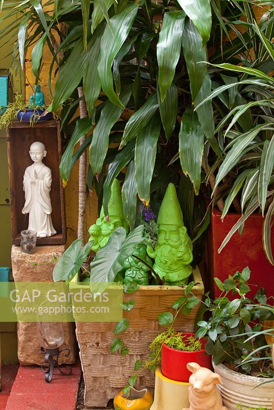 A collection of quirky pots and statues with two green garden gnomes and a white ceramic oriental figure next to a potted Dracaena fragrans.