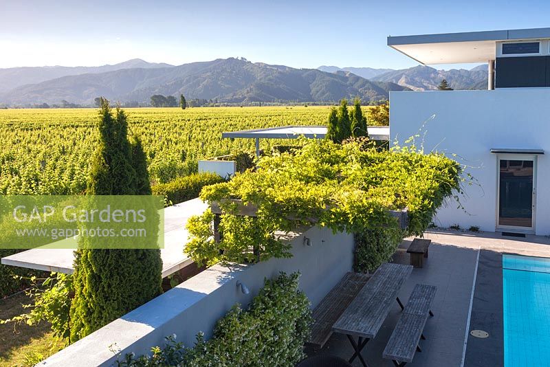 Vines covering a table and bench next to the pool  with views of the mountains and vineyards beyond at Bhudevi Estate garden, Marlborough, New Zealand.