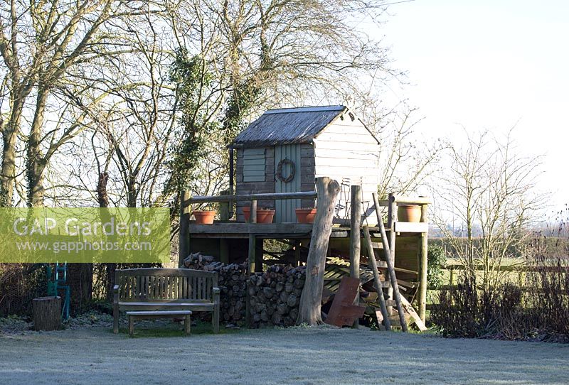 Child's play house on platform with wood store underneath. Wooden bench and table on a frosty morning. St Francis Cottage, Suffolk