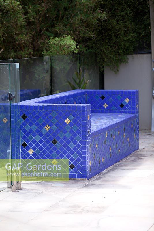 Rectangular Moroccan inspired blue tiled bench seat with gold and black accent tiles in front of a glass pool fence.