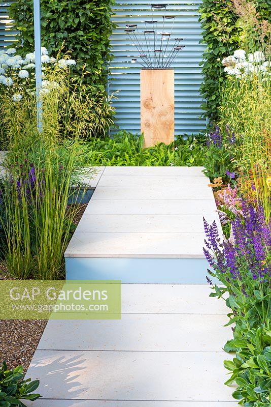 Retreat Garden, white paved pathway and step leading to wood and metal sculpture and steel louvred partitions. Planting is direct into the surrounding gravel with drought tolerant plants including stipa gigantea and salvias. Behind the step are ferns and chrysanthemums.  RHS Hampton Court Flower Show in 2016