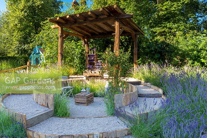 The Lavender Garden, view of garden with its rustic rough-hewn timber gazebo, approached by a circular, stepped gravel pathway. In the centre are chairs and a crate serving as a table. Lavenders include Lavandula angustifolia and L. x intermedia cultivars. RHS Hampton Court Flower Show in 2016