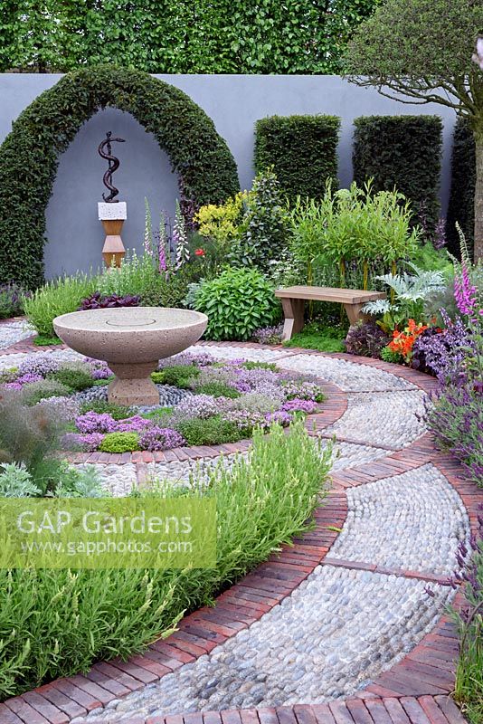 The St John's Hospice Garden, The Modern Apothecary.Brick and cobble stone path. Extensive planting of herbs around water basin in central circular bed. Oak seat in border and sculpture of Aesclepius on back wall. RHS chelsea Flower show 2016