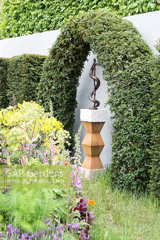 Sculpture of Asclepius's staff by Susan Bacon under an arch of Taxus baccata, Angelica archangelica in foreground - A Modern Apothecary, The St John's Hospice Garden, RHS Chelsea Flower Show 2016. Designer: Jekka McVicar, Sponsor: St John's Hospice