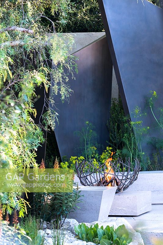 The Telegraph Garden, view of a fire bowl, irregular geometric bronze fin sculptures, seating area with stone benches surrounded by Isoplexus canariensis - Canary Island Foxgloves, Maytenus boaria, Schinus molle. RHS Chelsea Flower Show 2016. Designer: Andy Sturgeon - Sponsor: The Telegraph
