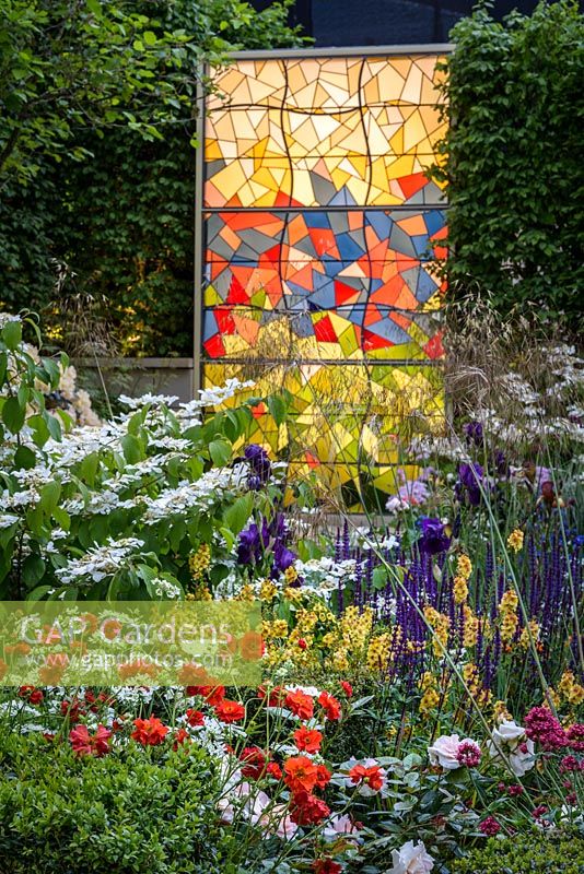 Evening in God's Own Country - A Garden for Yorkshire, with Geum, Salvias, Irises, Roses, Stipa gigantea, Viburnu 'Mariesii', lit by a modern stained window, inspired by York Minster. The RHS Chelsea Flower Show 2016. Designer: Matthew Wilson- Sponsor: Welcome ot Yorkshire - SILVER -PEOPLES CHOICE BEST SHOW GARDEN