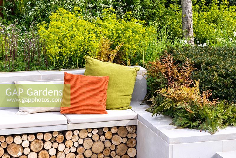 Detail of a bench with log store and colourful cushions surrounded by Dryopertis erythrosora, Euphorbia robbiae and Taxus baccata, The Sunken Retreat, RHS Malvern Spring Festival 2016. Design: Ann Walker, Graduate Gardeners
