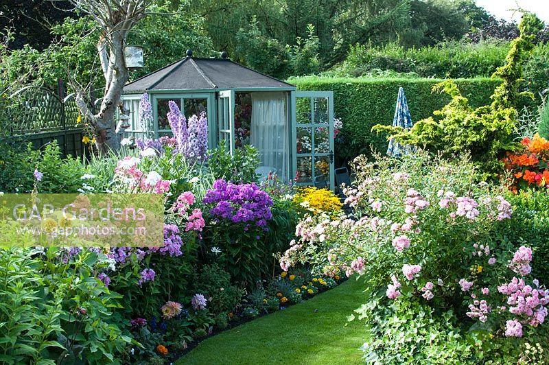 Colourful back garden with mixed borders filled with tender bedding plants, shrubs and perennials including Rosa 'The Fairy', Delphinium 'Sweet Sensation' and Phlox 'Elizabeth' 