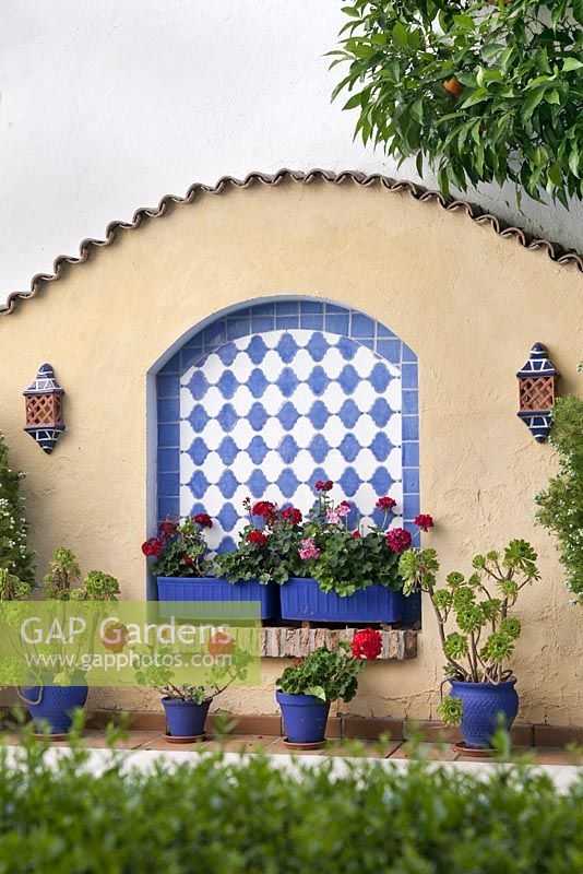 Red pelargoniums in blue painted terracotta pots in decorative tiled archway set into plastered wall feature - Cordoba, Spain