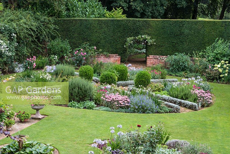 A circular parterre with pink and blue planting including Sedum 'Matrona', Caryopteris 'Heavenly Blue', Agapanthus 'Blue Moon', Cleome hassleriana 'Rose Deep', Knautia macedonica, Cuphea viscosissima and Gladiolus murielae. Behind, a yew hedge with entrance surrounded by climbing roses.