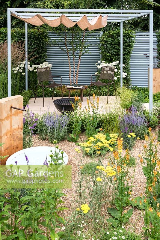 A enclosed contemporary town garden with stone patio seating area with fire pit under a steel pergola with retractable shade, water feature and a gravel bed planted with perennials and grasses. A Retreat Garden designed by Martin Royer. Hampton court 2016