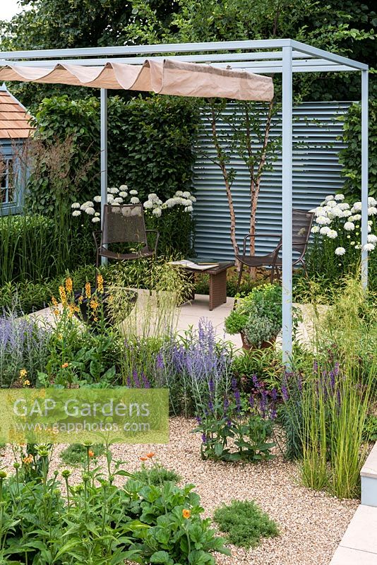 A enclosed contemporary town garden with stone patio seating area with fire pit under a steel pergola with retractable shade and a gravel bed planted with perennials and grasses. A Retreat Garden designed by Martin Royer. Hampton court flower show 2016 