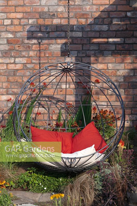 A contemporary spherical suspended metal chair against a red brick wall with perennials in hot colours -   July, Tatton Park RHS Flower Show 2014