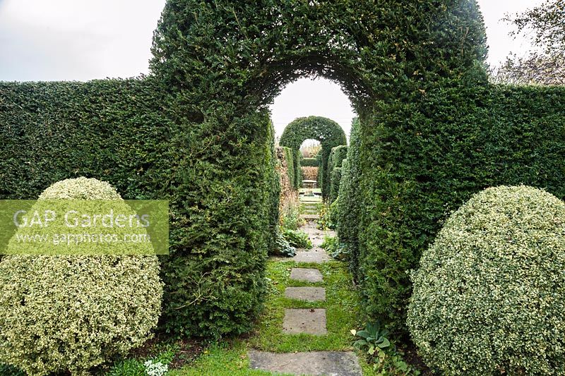 A clipped yew arch, marked by a pair of variegated box shrubs, frames a view through the garden between yew and hornbeam hedges to an urn, overflowing with water, sitting in a pond.