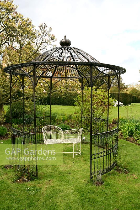 Elegant circular gazebo in black painted steel set in grass with a lovers seat beneath and recently planted climbers. 