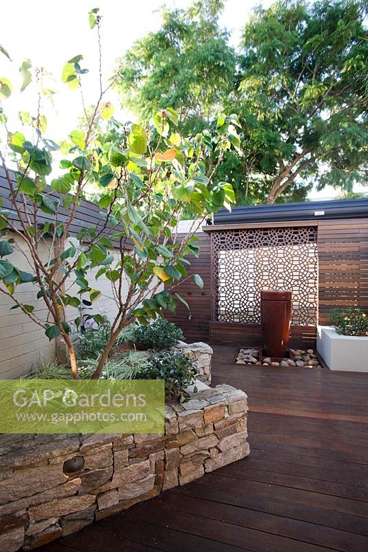 Inner city courtyard with featuring a raised bed with stone stack wall with a Cercis  tree,a freestanding ceramic water feature, a rusty corten decorative screen and a timber slat panel wall