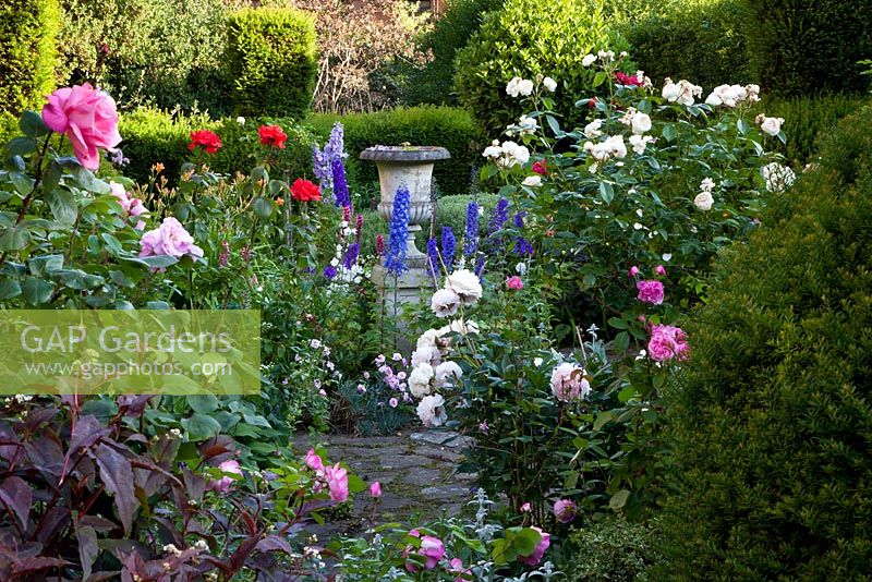View down The Bear Shop Gardens to a stone urn surrounded by Delphiniums, Dianthus, Rosa and Persicaria microcephala 'Red Dragon' 