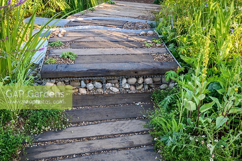 Timber and gravel path with gabion filled with stones as a step. The WWT Working Wetlands Garden. Hampton Court Flower Show 2016 - Designer: Jeni Cairn, Sponsor: Wildfowl and Wetland Trust supported by the HSBC Water Programme