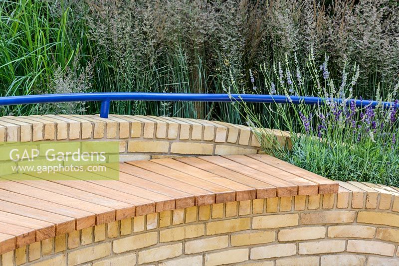 A curving brick bench with wood seating and blue curving handrail with Calamagrostis acutiflora 'Overdam', Lavandula 'Hidcote'. The Abbeyfield Society: a Breath of Fresh Air, RHS Hampton Court Palace Flower Show 2016. Design: Rae Wilkinson