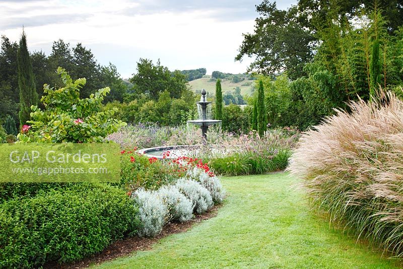 A curved grass path, bordered by clipped Cistus x loretii, Helichrysum italicum and Rosa 'Kadora' on the left and Miscanthus sinensis 'Autumn Light' on the right, leads to the Italian garden with a Florentine fountain in les Jardins de la Poterie Hillen.