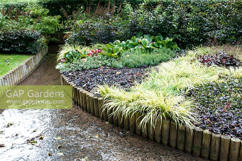 Raised bed above stream with mixed tapestry evergreen planting of Ajuga reptans 'Catlin's Giant' bugle Bergenia 'Eric Smith' Euphorbia cyparissias 'Fens Ruby' cypress spurge Hebe Heuchera Carex and Fagus sylvatica Purpurea hedge 