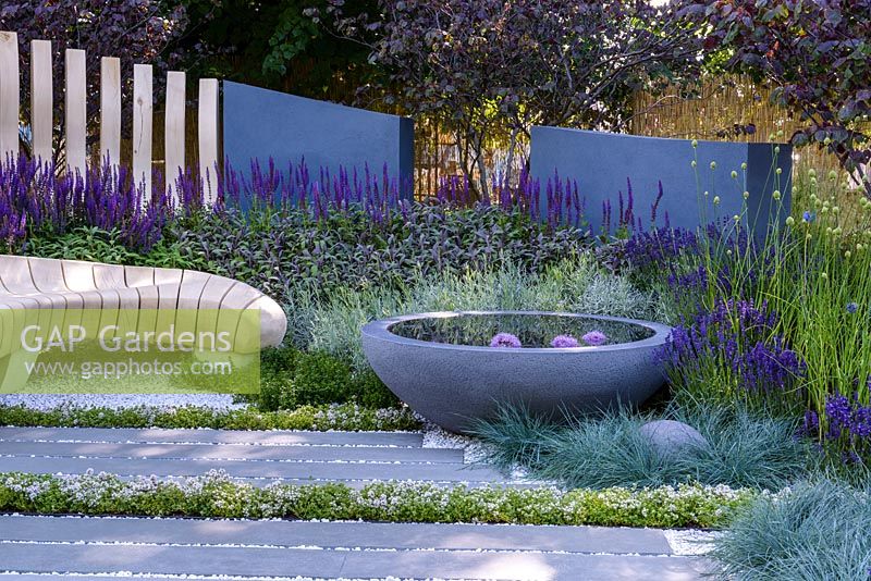 Gravel and stone patio with curved bleached oak wooden bench and fence, grey rendered wall, concrete containers and small pond. Planting includes Thyme and Rosemary, Salvia officinalis 'Purpurascens', Salvia tanzerin, Perovskia atriplicifolia 'Blue Spire'.  Living Landscapes: Healing Urban Garden, RHS Hampton Court Palace Flower Show 2015. Designer Rae Wilkinson