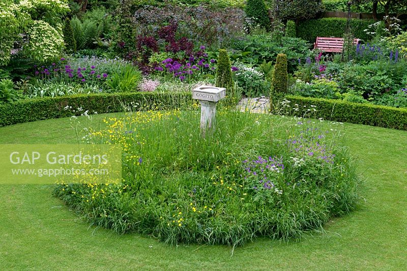 A cottage garden with circular lawn and raised wildflower mound planted with buttercups, cranesbill and cow parsley surrounding a bird bath. The formal structure is created by low box hedging and conical shaped yews.