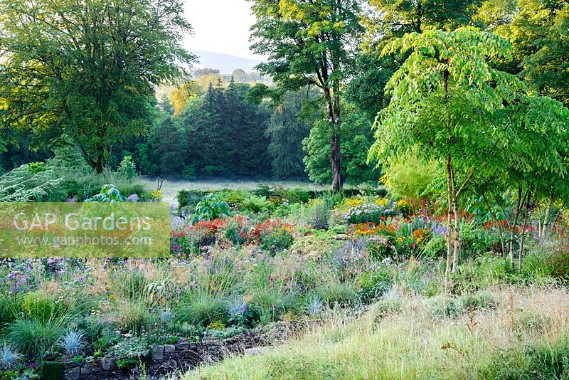 Beds planted with a matrix of herbaceous perennials and shrubs set between grassy slopes in the higher part of the garden in the foreground and cultivated farmland beyond with the Wicklow mountains glimpsed through a gap in the trees.