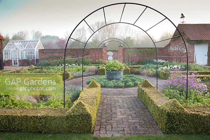 Spring beds in formal country garden with decorative metal arches, Buxus clipped hedge and Honesty, Sedum 'Autumn Joy', Wallflower, Narcissus and Tellima grandiflora. Garden: Ulting Wick, Essex, Owner: Philippa Burrough 