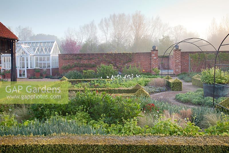 Spring beds in formal country garden with decorative metal arches, Buxus clipped hedge and Geranium, Sedum 'Autumn Joy', Narcissus, Tellima grandiflora and Stipa. Garden: Ulting Wick, Essex, Owner: Philippa Burrough
