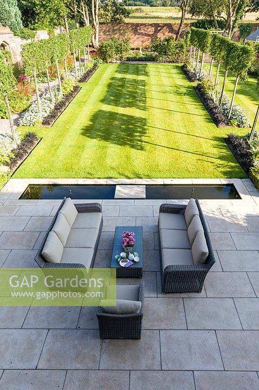 An elevated view of the newly contemporary English garden in Cheshire, designed by Louise Harrison-Holland and photographed in August. The patio area, with garden furniture and rill lead into a lawned walled garden, with plants including pleached Pyrus calleryana 'Chanticleer', Japanese anemones, Stachys, Persicaria, Ophiopogon planiscapus 'Nigrescens' and Miscanthus sinensis 'Kleine Fontaine'.