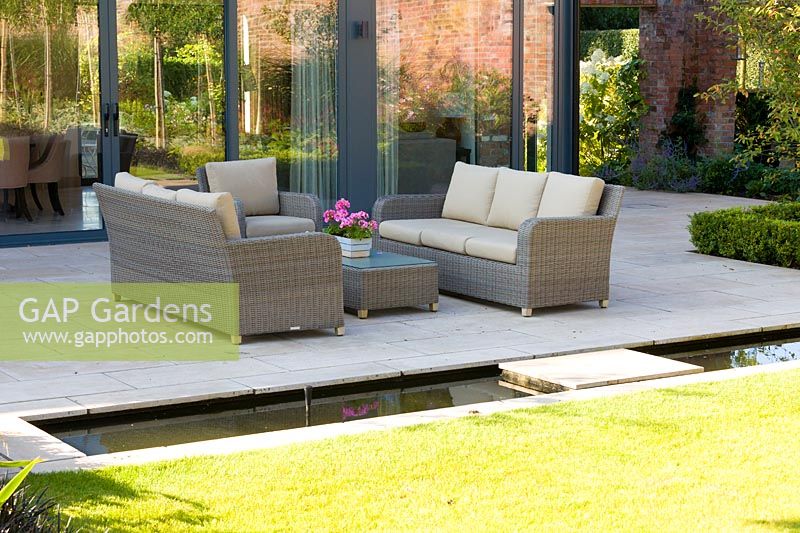 A patio area, with seating and a rill, in a modern Cheshire country garden, designed by Louise Harrison-Holland. Planting includes box hedging and a Pelargonium in a pot