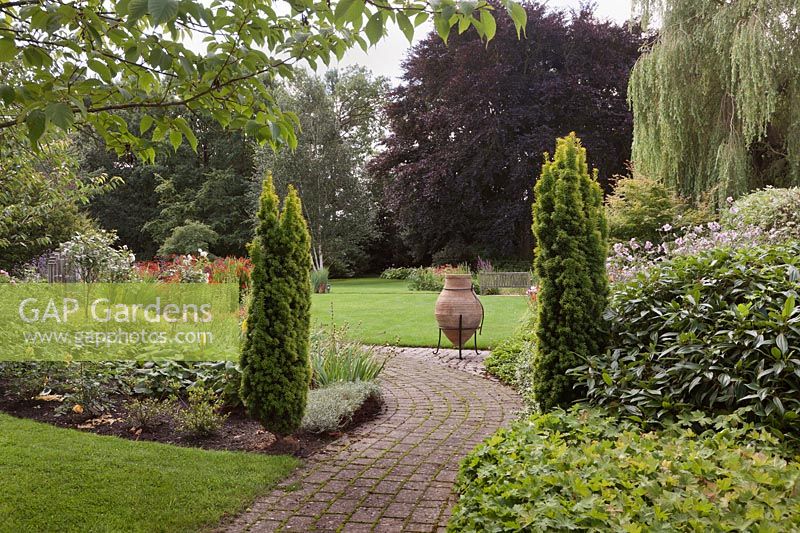 A curving brick path between flower beds with lawns, mature trees and terracotta urn on stand 