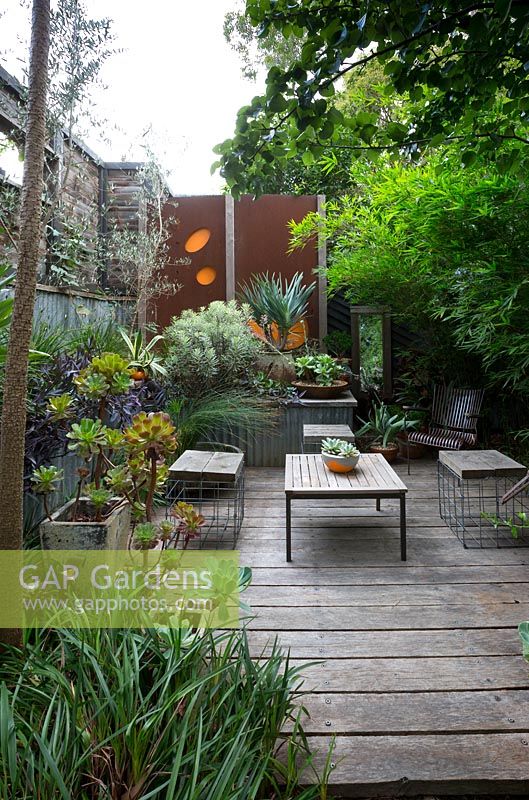 A sunken outdoor entertaining area made from timber sleepers with table and chairs, a heavily planted raised garden bed and decorative corten steel screens.