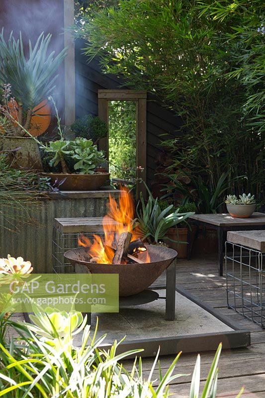 A sunken fire pit area with a freestanding metal fire pit, on a timber deck surrounded by raised garden beds and rusty corten steel screens.