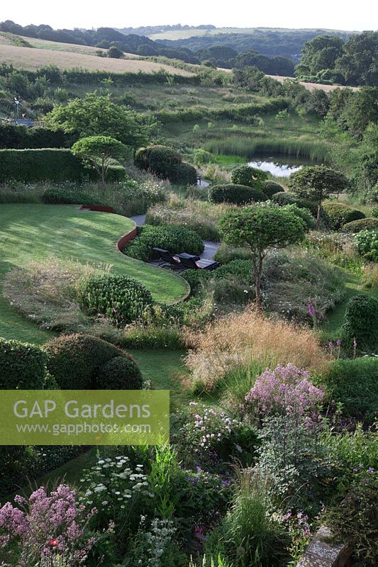 Overview of garden with countryside views beyond. Curving rusted Corten steel walls, Pinus mugo, clipped Crataegus x lavalleei 'Carrierei', Taxus baccata - Yew topiary, meadows and  natural pond. Borders with Stipa gigantea, Cenolophium denudatum, Eryngium giganteum, Thalictrum 'Elin' and Rosa 'Rosemoor'.
