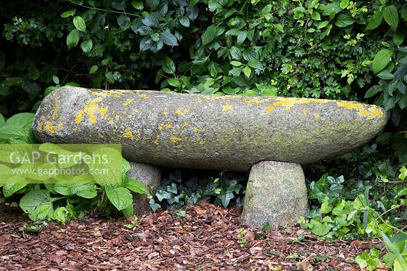A stone bench covered in Lichen in a shady corner with bark mulch