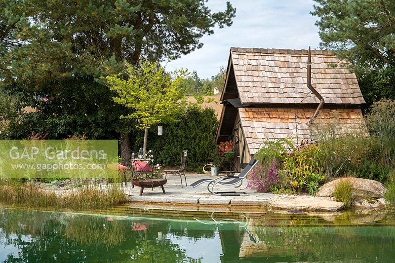 There is a wide rest area with fire bowl and deck chair next to two-storey witches' cottage. In the foreground, the natural swimming pond. Plants include Pinus sylvestris, Styrax japonica and Lespedeza thunbergii