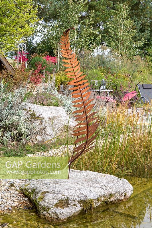 Next to the shallow water zone a metal sculptural fern on a granite boulder. In the background a rest area, birches and perennial planting