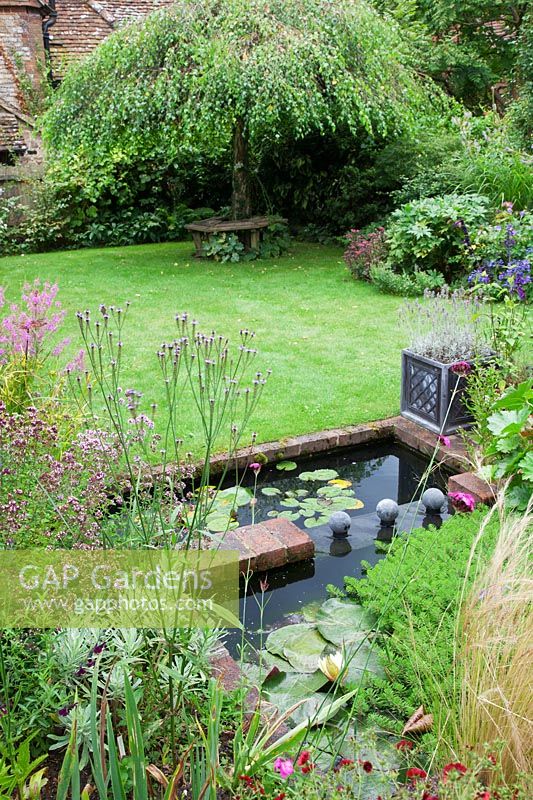 A lead covered water feature two tier pond with water lily foliage in a small low maintenance modern cottage garden. Planting includes Stipa tenuissima, Verbena hastata. Grass lawn leads to the weeping Birch - Betula pendula 'Youngii' with rustic wooden tree seat.
