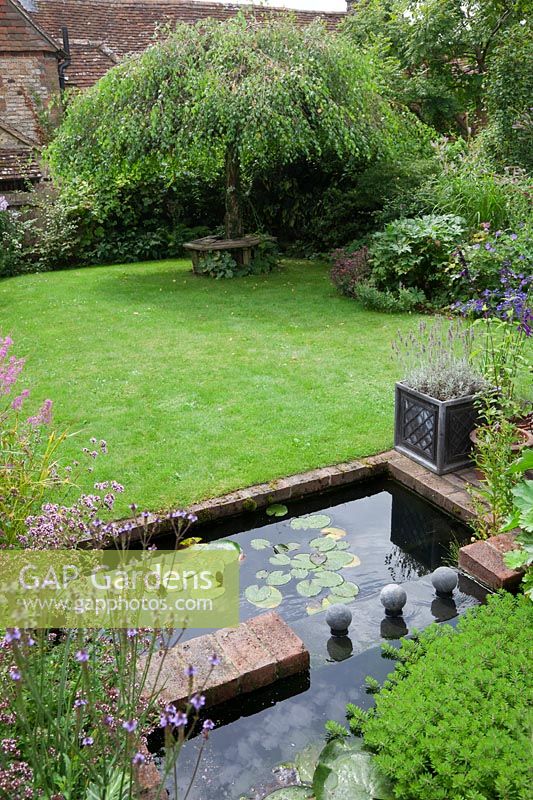 A lead covered water feature two tier pond with water lily foliage in a small low maintenance modern cottage garden. Planting includes Verbena hastata, a grass lawn leads  to the weeping Birch - Betula pendula 'Youngii' with rustic wooden tree seat.