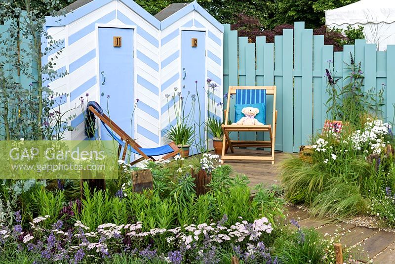 Blue and white painted beach huts and deck chairs on decking, driftwood posts, gravel, pebbles and planting of grasses, Eucalyptus gunnii 'Azura' underplanted with Achillea ptarmica 'The Pearl', Achillea millefolium 'Wonderful Wampee', Nepeta racemosa 'Walker's Low', Verbena bonariensis, Stipa tenuissima in front of blue fence - By The Sea - RHS Hampton Court Palace Flower Show 2017 - Design: James Callicott - Sponsor: Southend Borough Council.