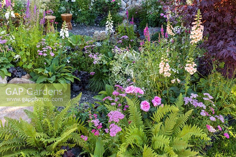 Carved timber seats in gravel area surrounded by deep reds, cream and pink colour themed planting with Digitalis, Stipa gigantea and Roses - Great Gardens of the USA- The Oregon Garden, RHS Hampton Court Palace Flower Show 2017- Designer: Sadie May Stowell