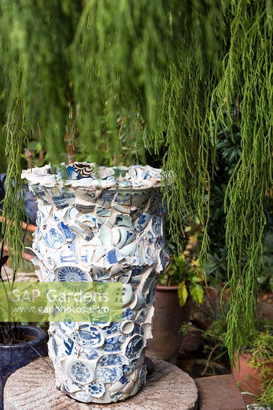 Large urn faced with pieces of broken blue and white china surrounded by a weeping juniper. Sculptor and ceramicist Marcia Donahue's garden in Berkeley, California.