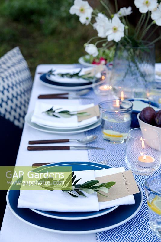 Outdoor dining table dressed in shades of blue and with a vase of Japanese anemones. Pockets are made in the folded napkins to put name card in and each setting is finished with an olive branch. Tealights in glasses light the table as dusk falls