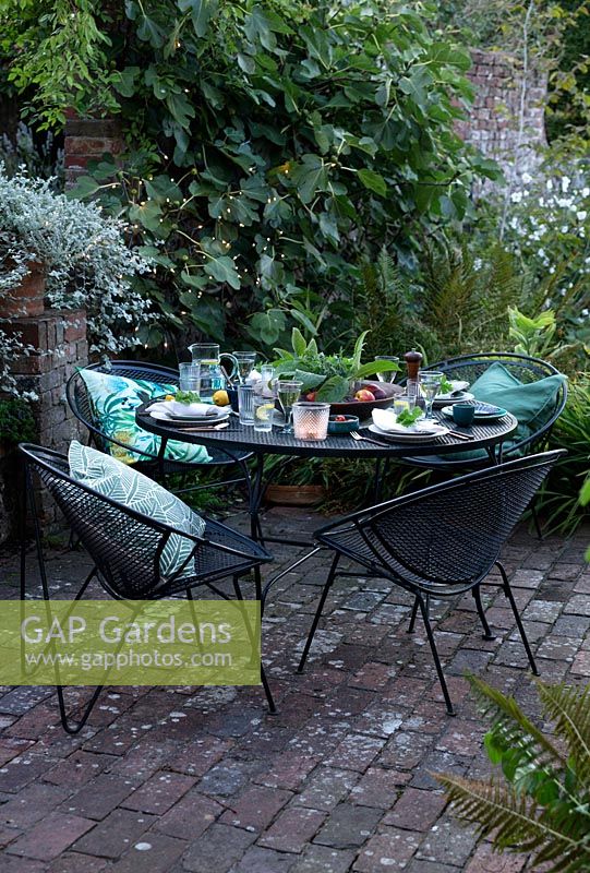 Outdoor dining area under a fig tree on a brick patio. Table laid with greens and fresh whites, napkins dressed with a geranium leaf and the centre of the table has a wooden bowl of fresh green leaves from the garden surrounded by fruit
