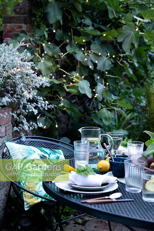 Dusk falls on the outdoor dining area under a fig tree. Fairy lights are in the trees creating a cosy setting and light up the area. Table laid with greens and fresh whites and napkins dressed with a geranium leaf