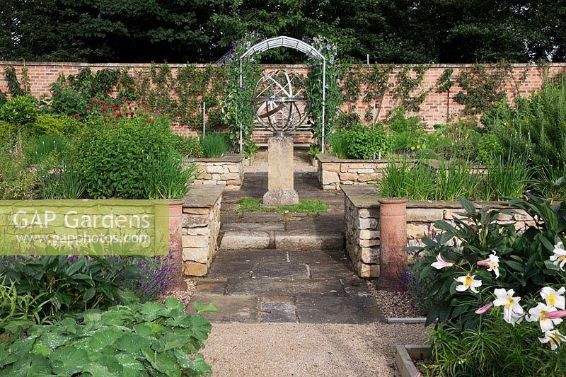 The herb garden in walled kitchen garden with focal point Armillary Sphere by David Harber. Raised beds with herbs including Rosemary, Monarda, Mint and Chives, made of drystone walls surrounded by Lavandula 'Imperial Gem'.   Steel arches with Sweet Peas and Flower beds with Lilies and Alchemilla mollis. Decorative chimney pots.