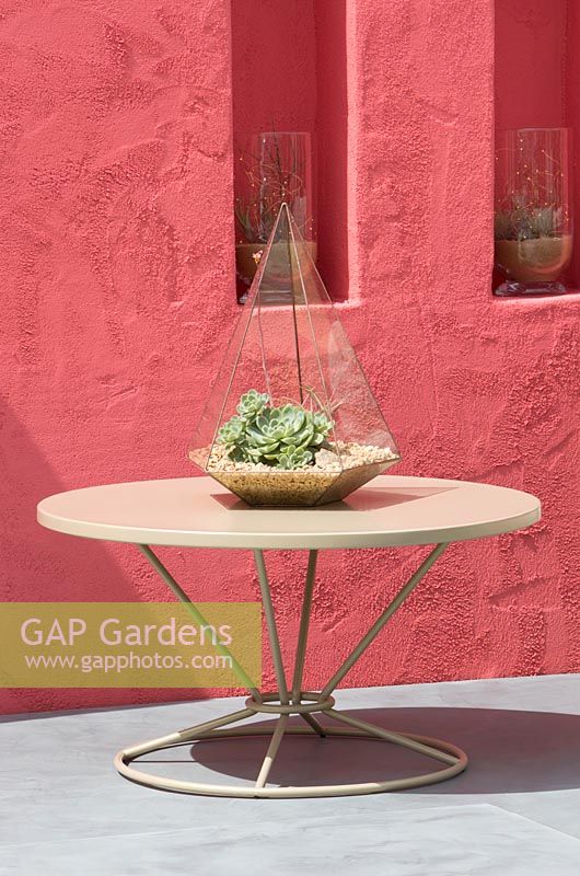 Beneath A Mexican Sky Garden - View of patio table with a terrarium with succulent, rendered red wall with alcoves and plants growing in glass vases - RHS Chelsea Flower Show 2017