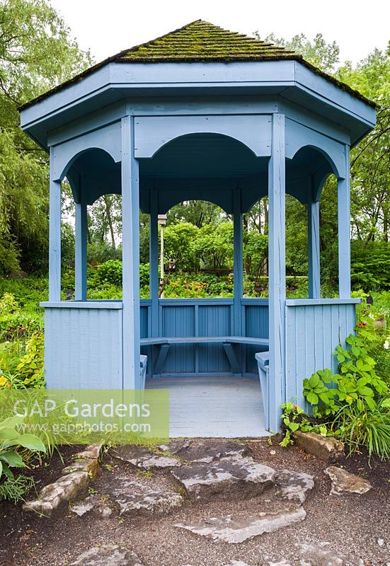 Blue painted wooden gazebo with built-in sitting benches and cedar shingles covered in green Bryophyta - Moss overlooking pond in summer, Centre de la Nature public garden, Saint-Vincent-de-Paul, Laval, Quebec, Canada